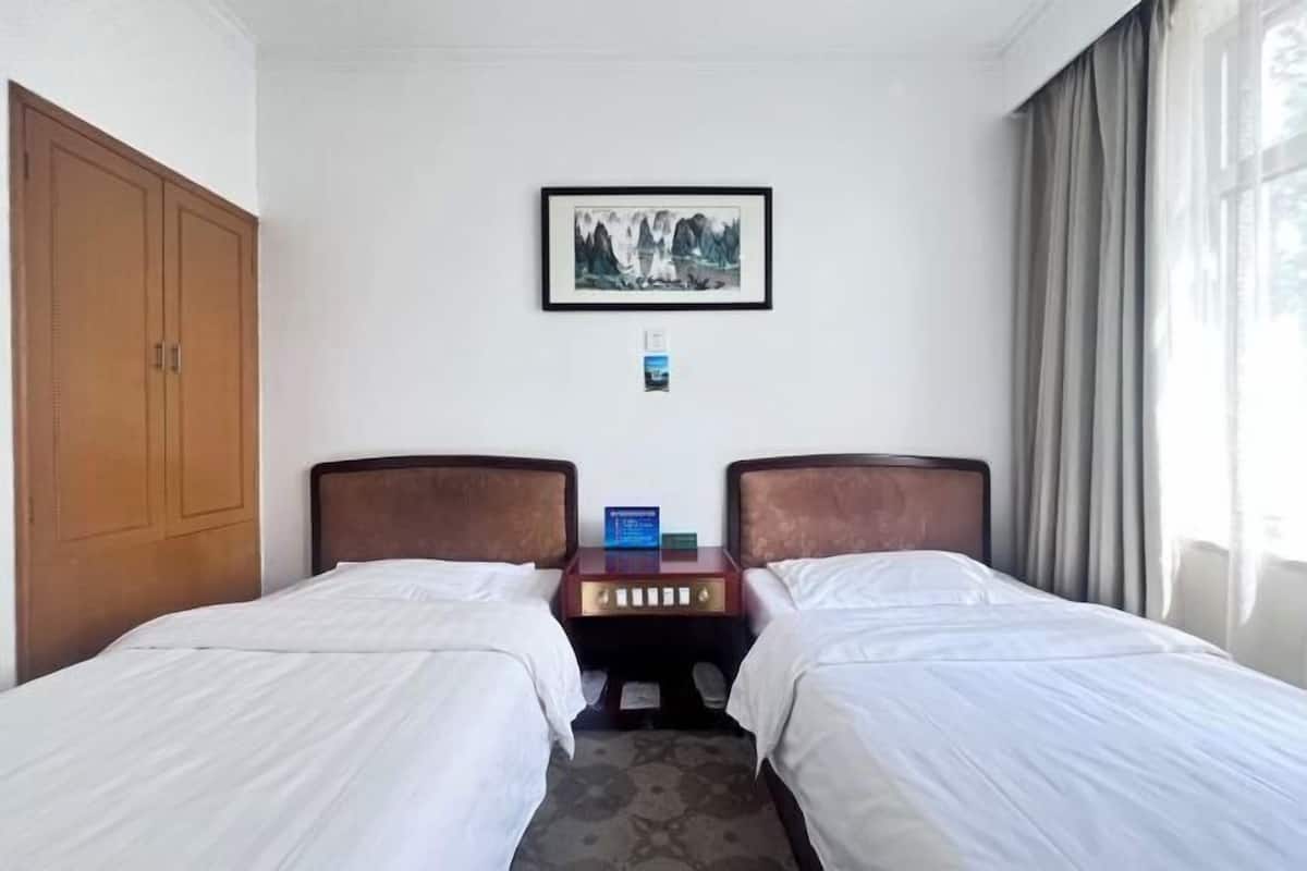 A hotel room with two beds, a wooden wardrobe, a framed picture on the wall, and a bedside table with a lamp.