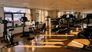 Uma academia ampla com várias esteiras, bicicletas ergométricas e equipamentos de ginástica. Grandes janelas oferecem luz natural e vista para o exterior.