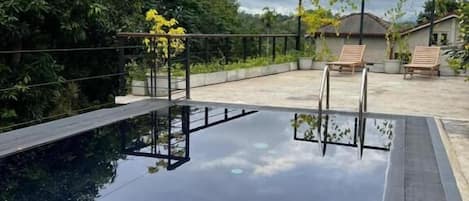Una piscina infinita con cubierta de madera y sillones de descanso, rodeada de exuberante vegetación y un cielo nublado.