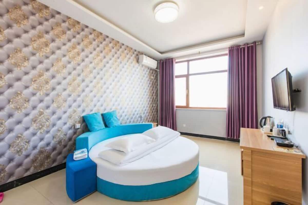 Quarto de hotel com cabeceira estofada, cama redonda com roupa de cama azul, escrivaninha de madeira e televisão na parede.