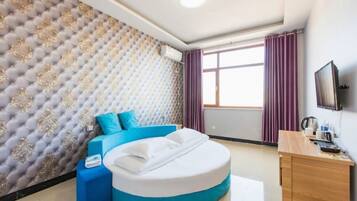 Una habitación de hotel con cabecera capitonada, cama redonda con ropa de cama azul, escritorio de madera y televisor en la pared.