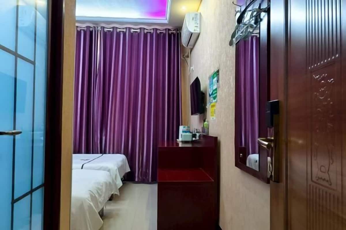 Quarto de hotel com duas camas, cortinas roxas, uma pequena escrivaninha com espelho e ar condicionado fixado na parede.