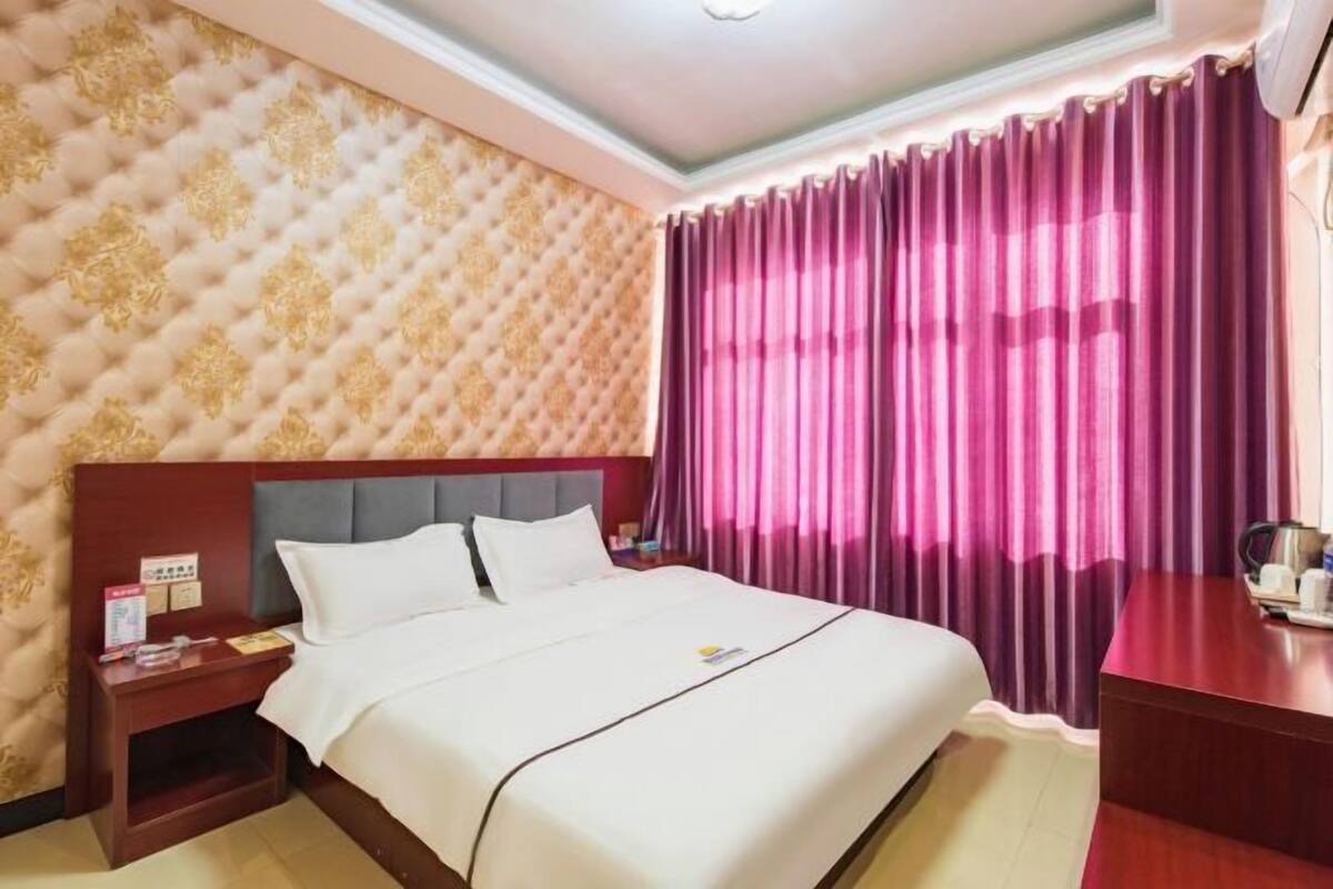 Quarto de hotel com cama, mesa de cabeceira, escrivaninha de madeira e janela com cortinas roxas.