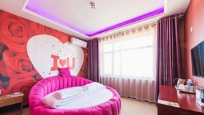 A room with a round, pink bed, a large heart-shaped decoration on the wall, a desk with a TV, and a window with curtains. - jujia lianhe jiudian (Boye)