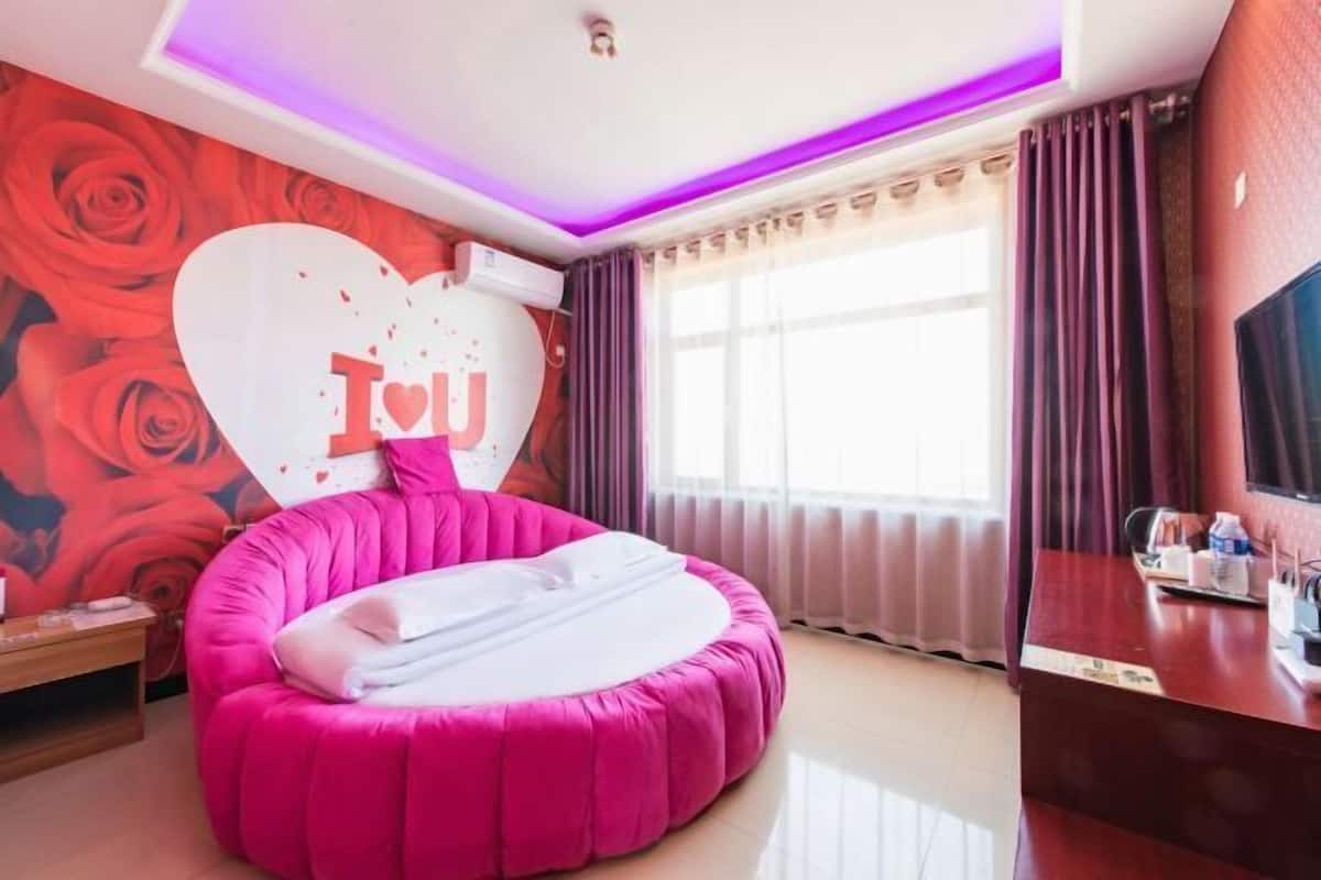 Quarto com cama redonda rosa, grande decoração em formato de coração na parede, escrivaninha com televisão e janela com cortinas.