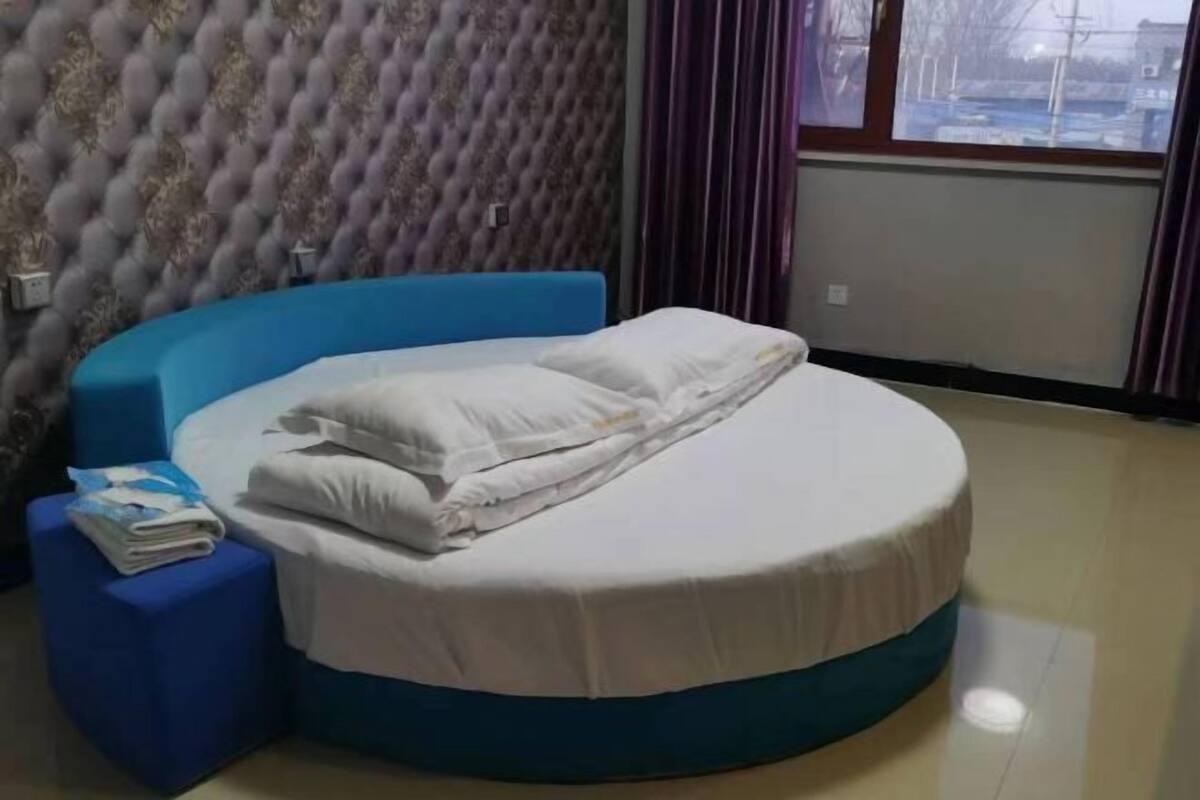 Uma cama redonda com cabeceira azul em um quarto com papel de parede estampado e uma janela com cortinas.