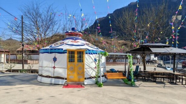 A traditional yurt with a yellow door, surrounded by a concrete area with benches and a small structure with a roof. - Xinglong Xiaoshan Farmhouse (Xinglong)