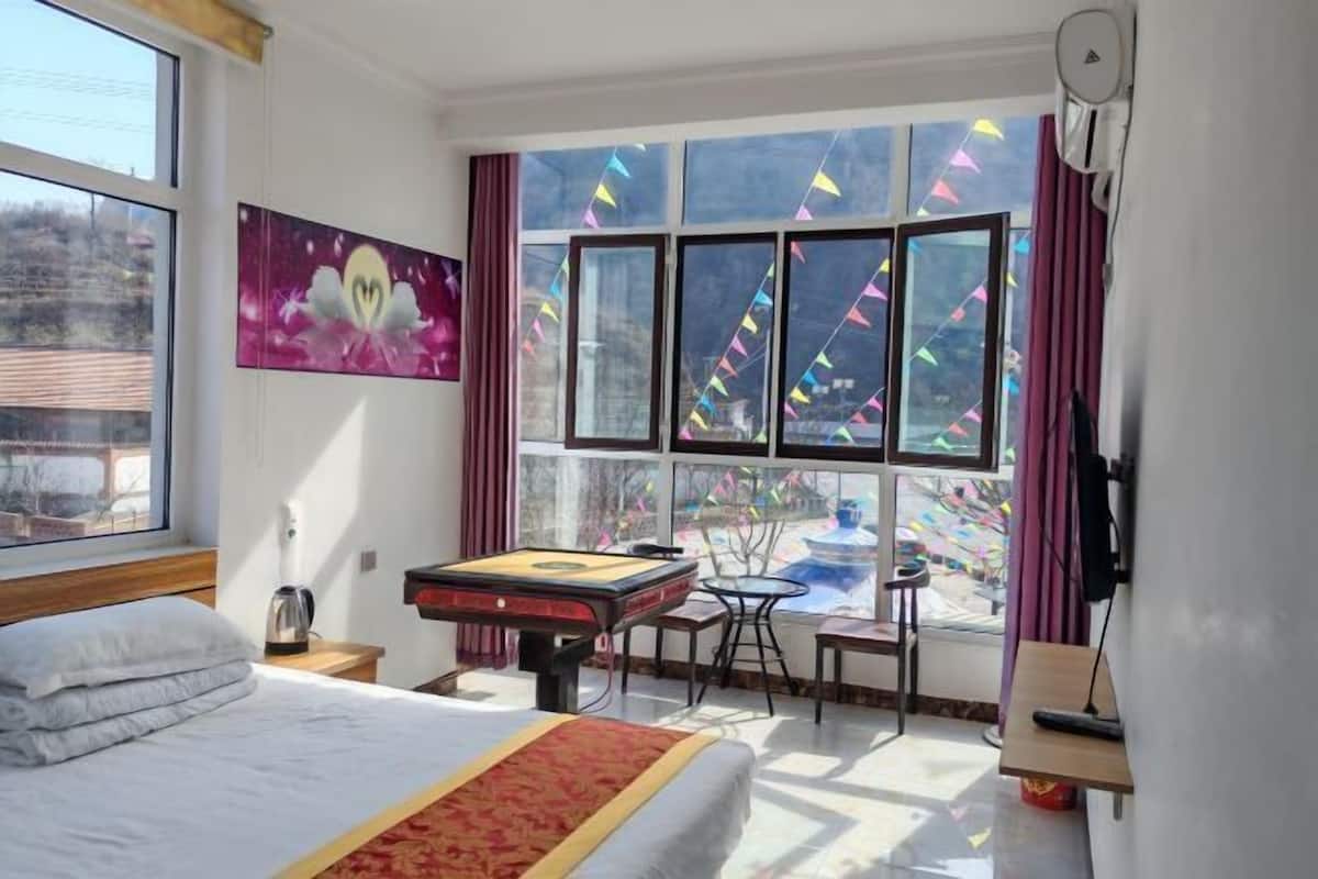 A hotel room with a bed, a desk, a table, a window with curtains, and a painting on the wall.