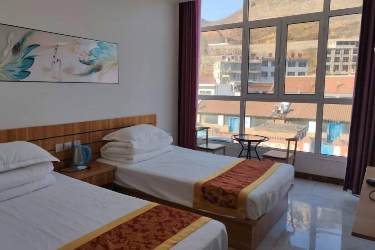 A hotel room with two beds, a painting on the wall, a window with a view of buildings, and a small table with chairs.