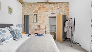 Une chambre avec un mur en pierre, un lit agrémenté d’oreillers à motifs, un radiateur et une porte en bois donnant sur une autre pièce.