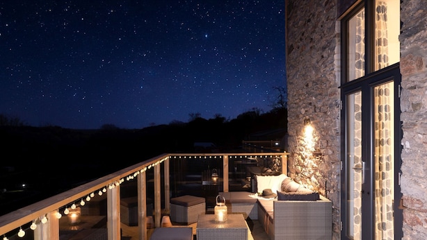 Un espace extérieur aménagé avec des meubles en osier et des guirlandes lumineuses, adossé à un mur de pierre, sous un ciel étoilé.