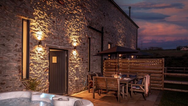 A stone building with a hot tub, wooden outdoor seating, and string lights. - One of 3 cottages, this beautiful barn conversion with a hot tub and EV charger is the perfect place (Paignton)