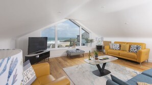 Ein modernes Wohnzimmer mit großem Fenster mit Blick auf einen Strand, einem gelben Sofa, einem Glastisch und einem Flachbildfernseher.