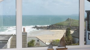 Blick durch ein Fenster auf die Küste, mit Sandstrand, felsigen Klippen und einem kleinen Hügel mit einer Bebauung.