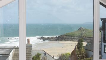 A coastal view through a window, featuring a sandy beach, rocky cliffs, and a small hill with a structure on top.