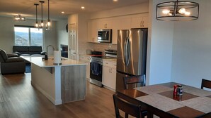 A modern kitchen with stainless steel appliances, a dining area with a wooden table, and a view of the outdoors through a large window. - Home in Fort Saskatchewan (Fort Saskatchewan)