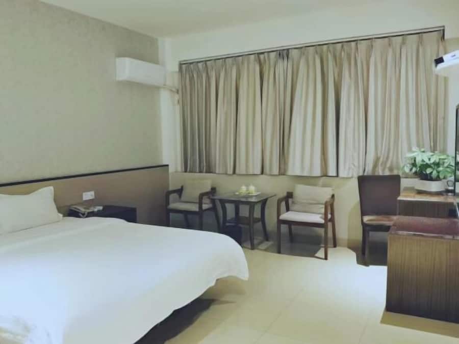 A hotel room with a large bed, a small table with chairs, a desk with a chair, and a window with curtains.