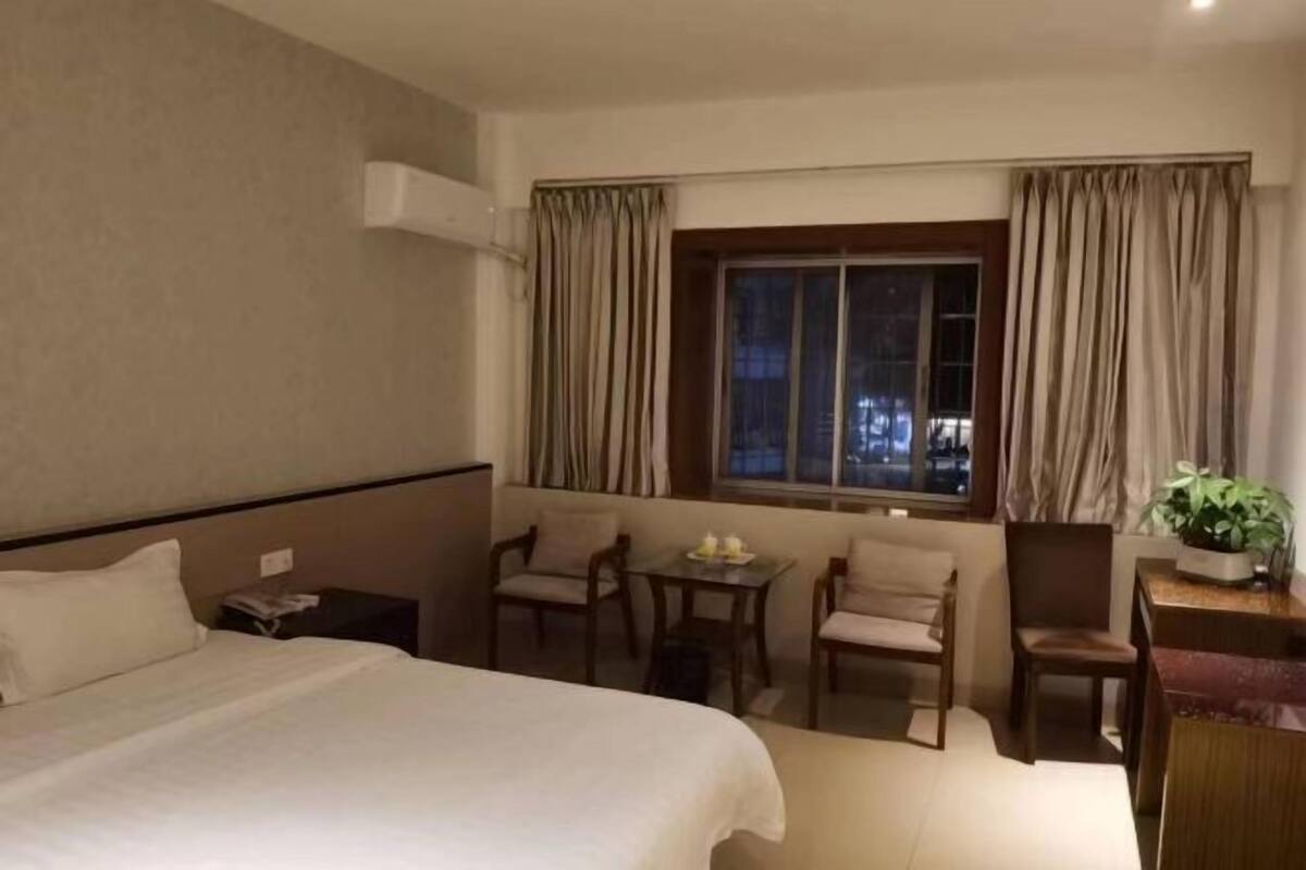 Une chambre d’hôtel avec un lit, une petite table et des chaises, une fenêtre avec des rideaux et un bureau agrémenté d’une plante.