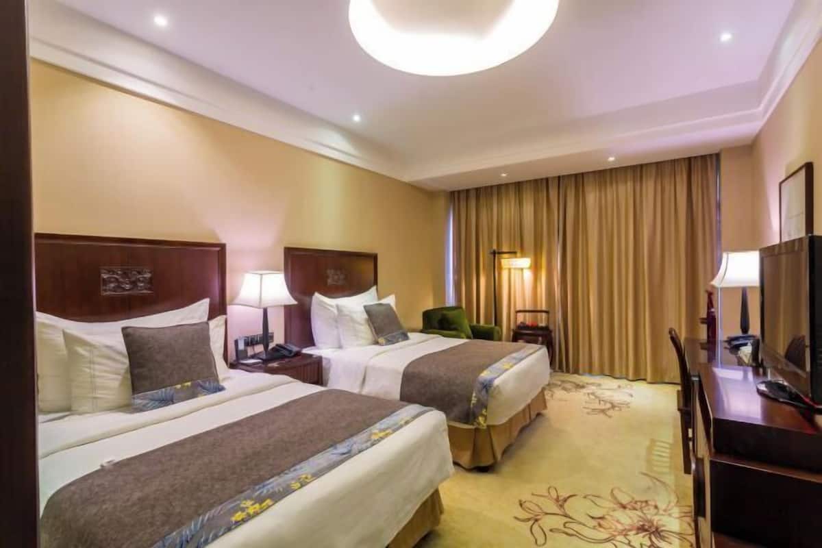 Een hotelkamer met twee bedden, een bureau, een stoel, een lamp en een groot raam met gordijnen.