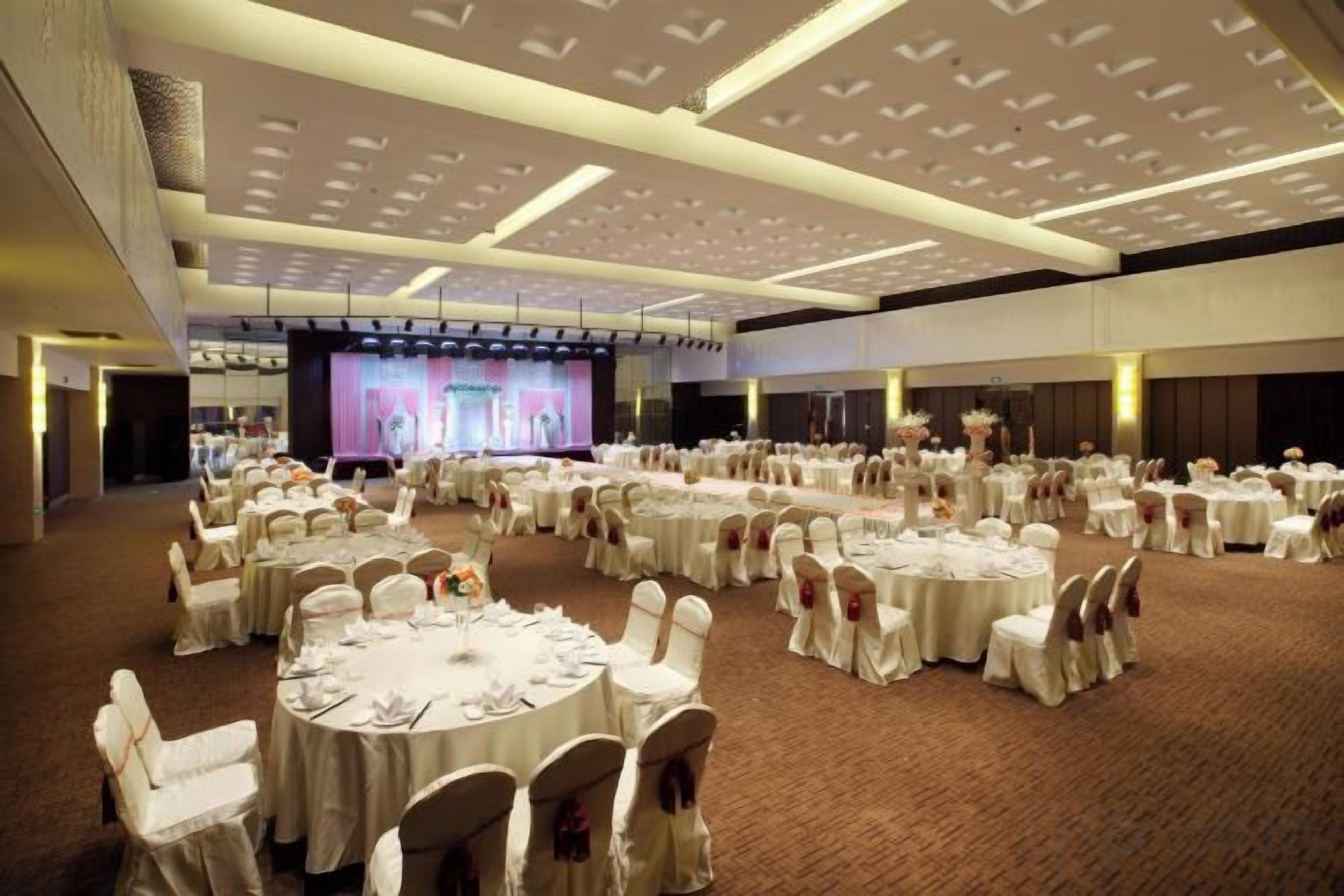 Un amplio salón de banquetes con mesas redondas dispuestas para eventos, un escenario con una pantalla y un techo alto con iluminación empotrada.