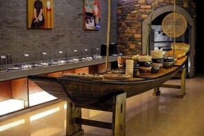 A long wooden table with food items and a woven basket, set against a brick wall with artwork.