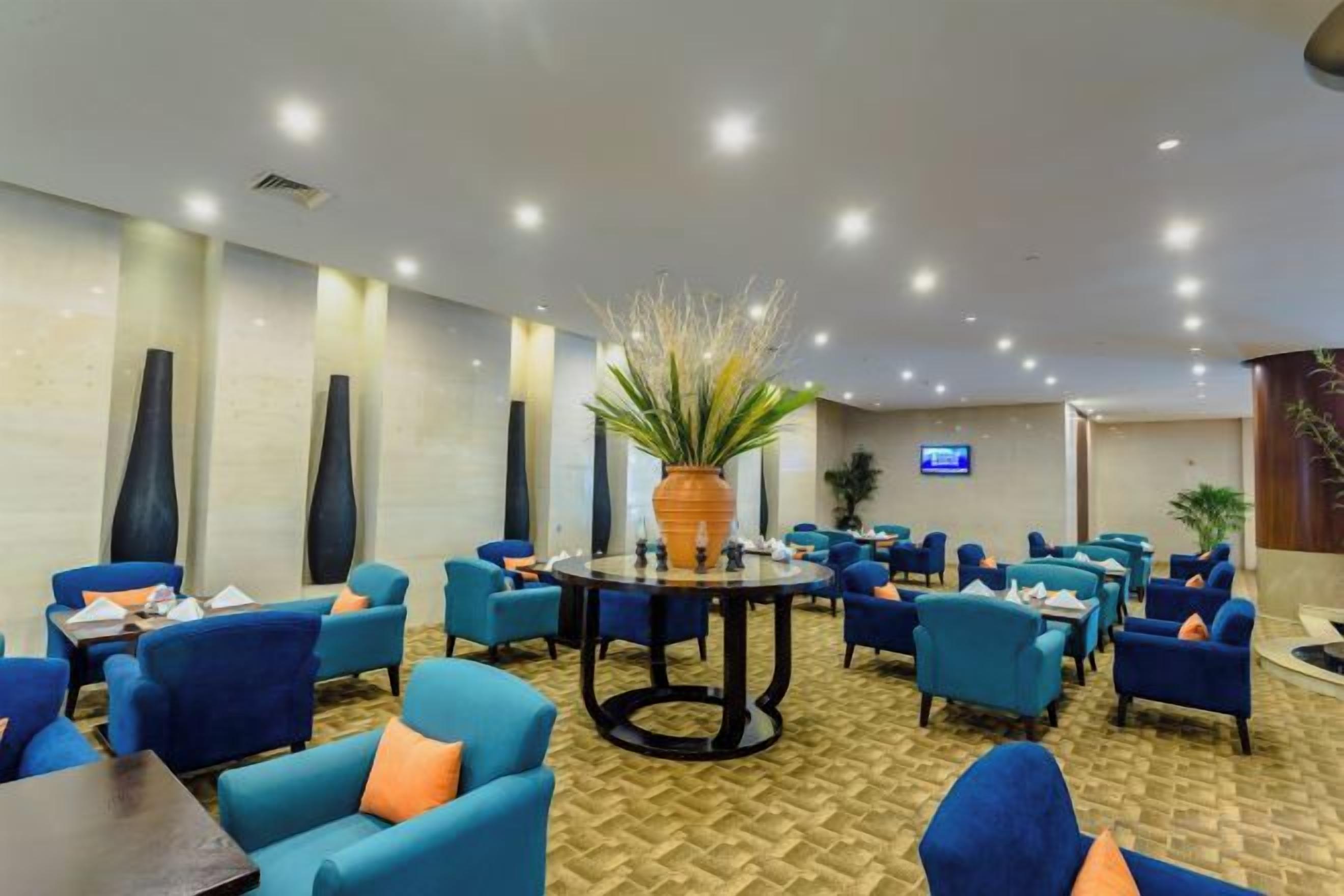 Un hall d’hôtel avec des tables rondes, des fauteuils bleus et une grande plante en pot.
