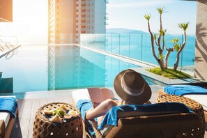 Une personne se détend au bord d’une piscine, un livre à la main, et profite de la vue sur l’eau d’un bleu limpide et sur la mer.