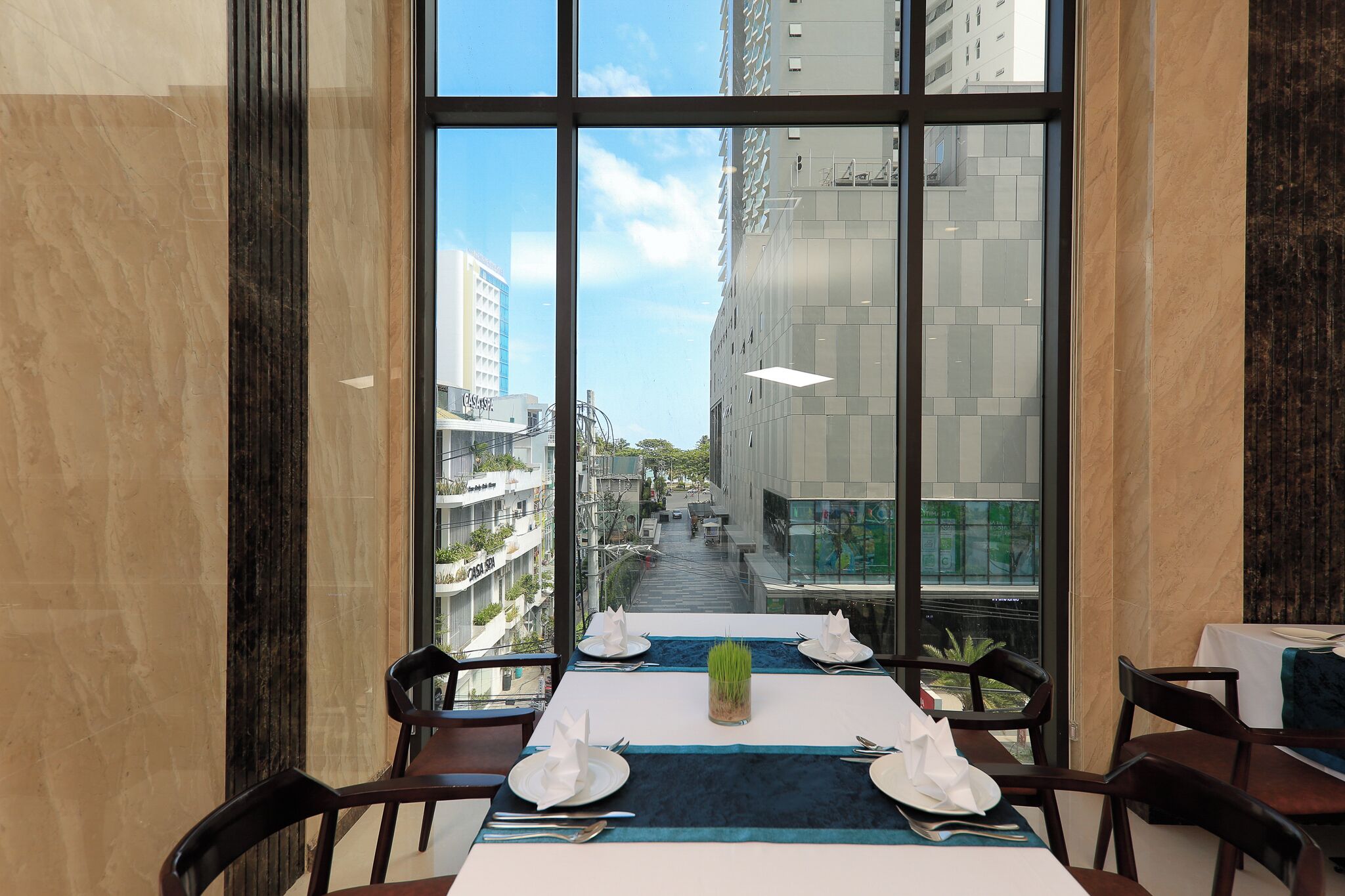 A dining area with a table set for four, a vase of green plants, and a view of a cityscape through large windows.