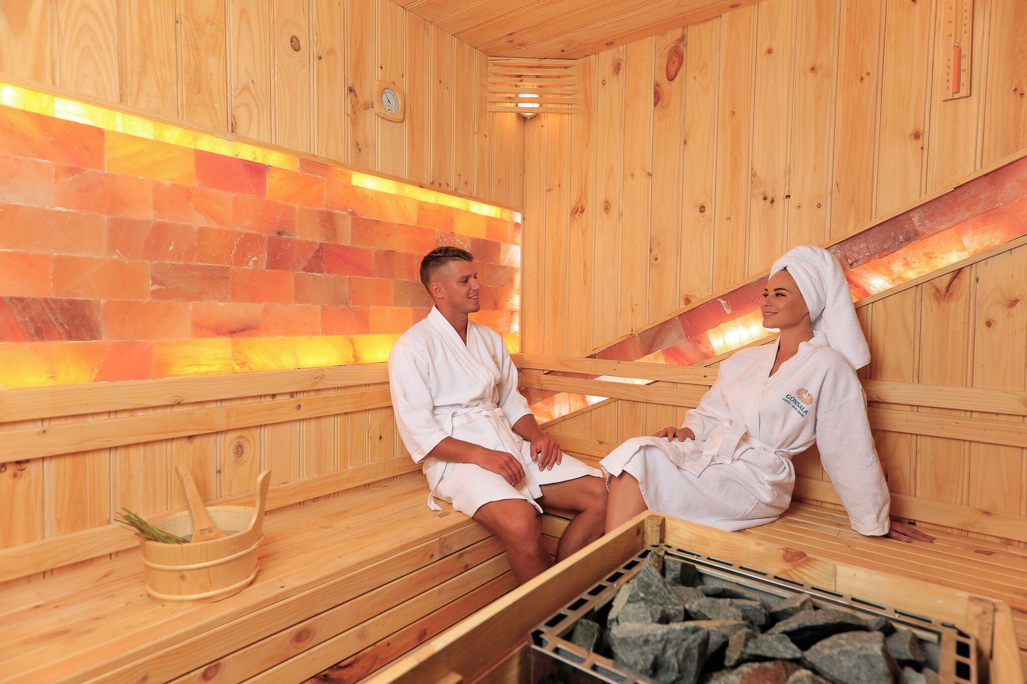 Dos personas en una sauna con paredes de madera y un revestimiento de azulejos al fondo.