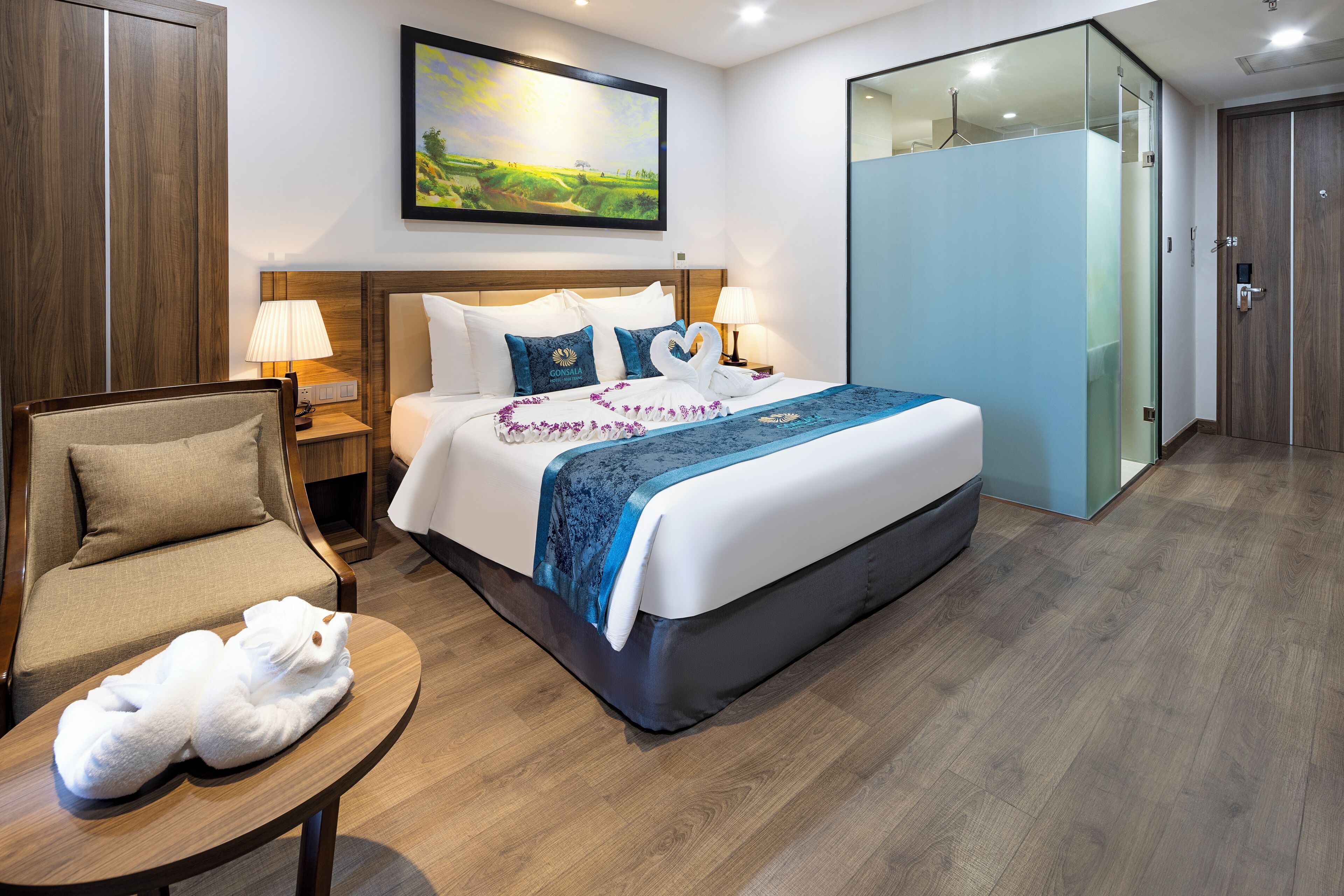 A hotel room with a bed, a chair, a table with towels, a nightstand, a lamp, a painting, and a wardrobe.