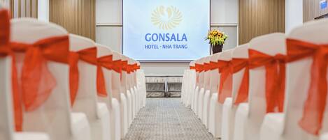 A hotel interior with rows of white chairs with red sashes, a projection screen displaying the hotel name, and a floral arrangement on the right.