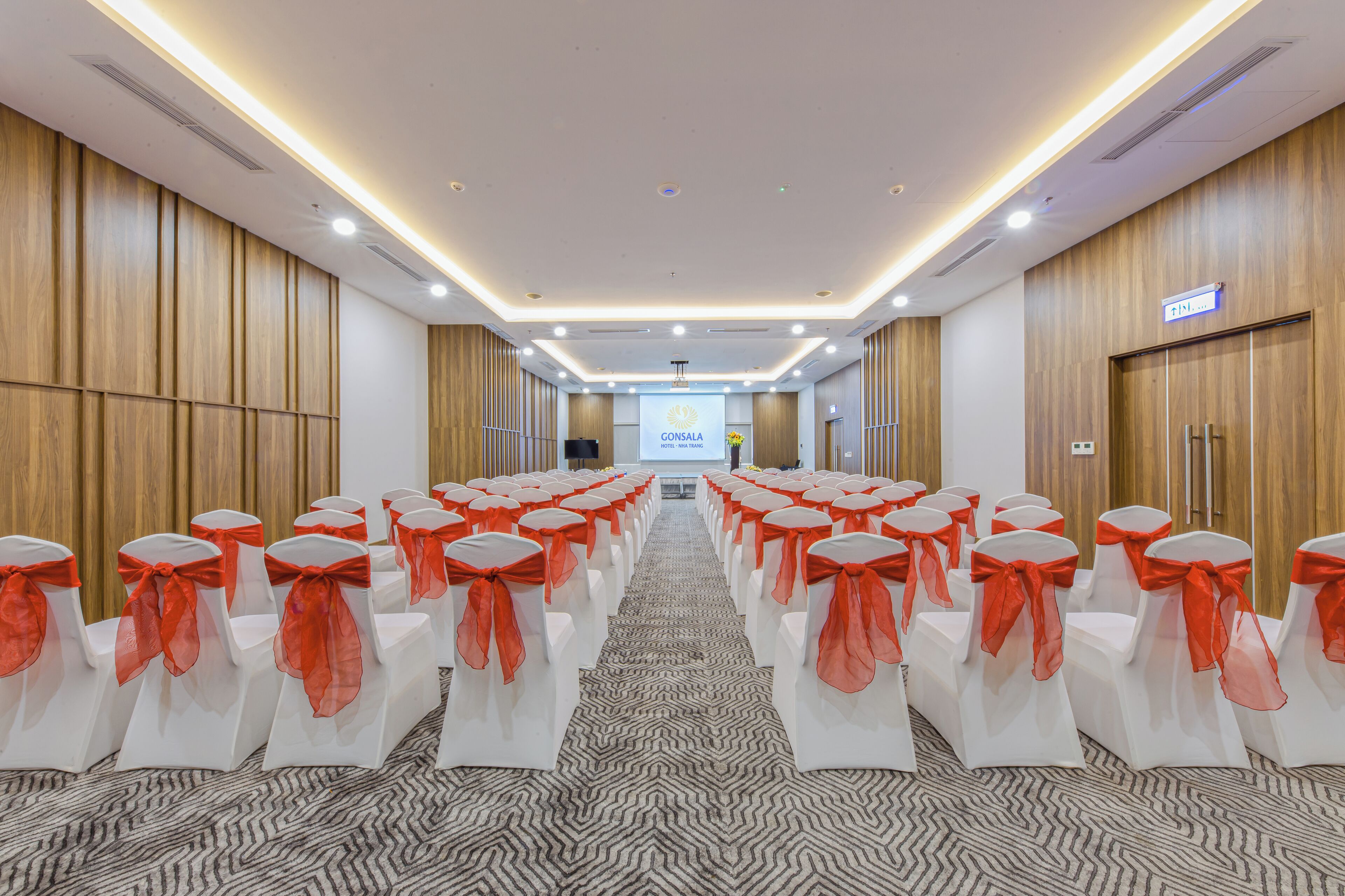 A conference room with rows of white chairs with orange sashes, wooden panel walls, and a carpeted floor.