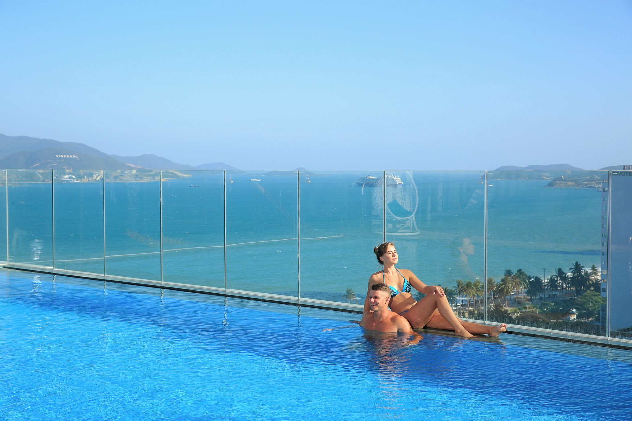 A couple relaxing by a pool with a view of the ocean and distant hills.