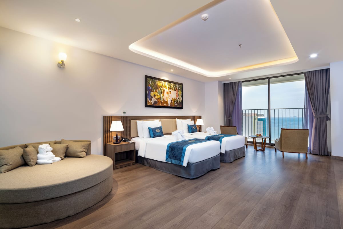 Ein Hotelzimmer mit gebogenem Sofa, zwei Betten mit passenden Kopfteilen, einem Bild an der Wand, einem Balkon mit Vorhängen und Blick aufs Meer.