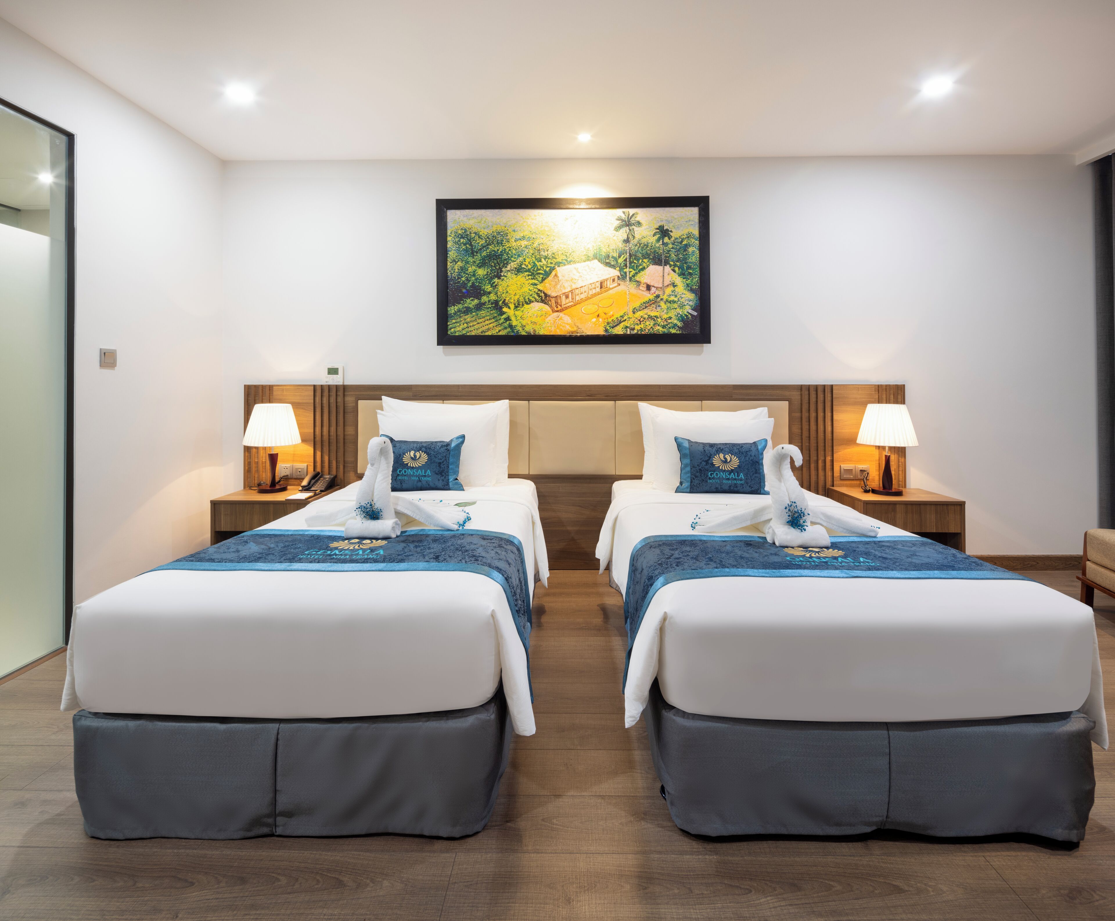 A hotel room with two beds, each with a matching pillow and bedside table. A painting is on the wall above the beds.