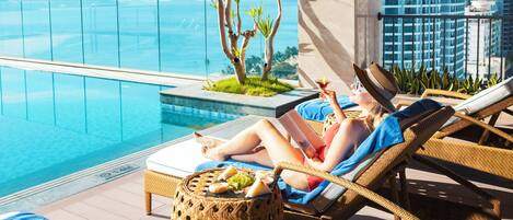 Zona de descanso junto a la piscina con vistas al océano y al horizonte de la ciudad, con sillones de mimbre, una pequeña mesa con comida y una barrera de vidrio.