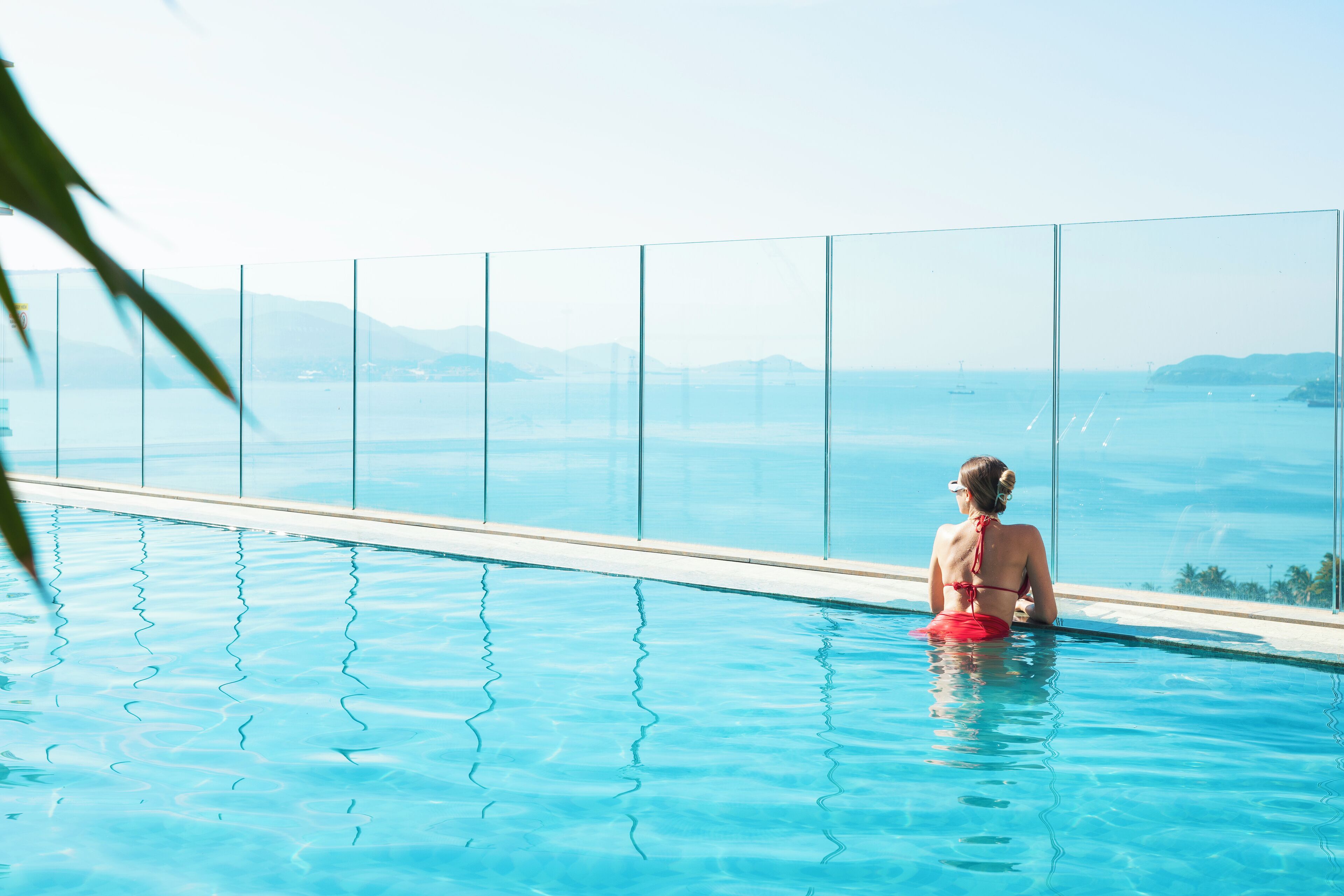 A person sitting in a clear glass-bottomed infinity pool with a view of the sea and distant mountains.