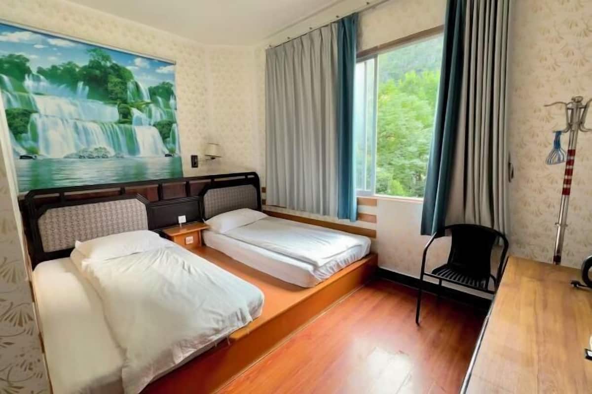 A hotel room with two beds, a desk, a chair, a window with curtains, and a painting of a waterfall.