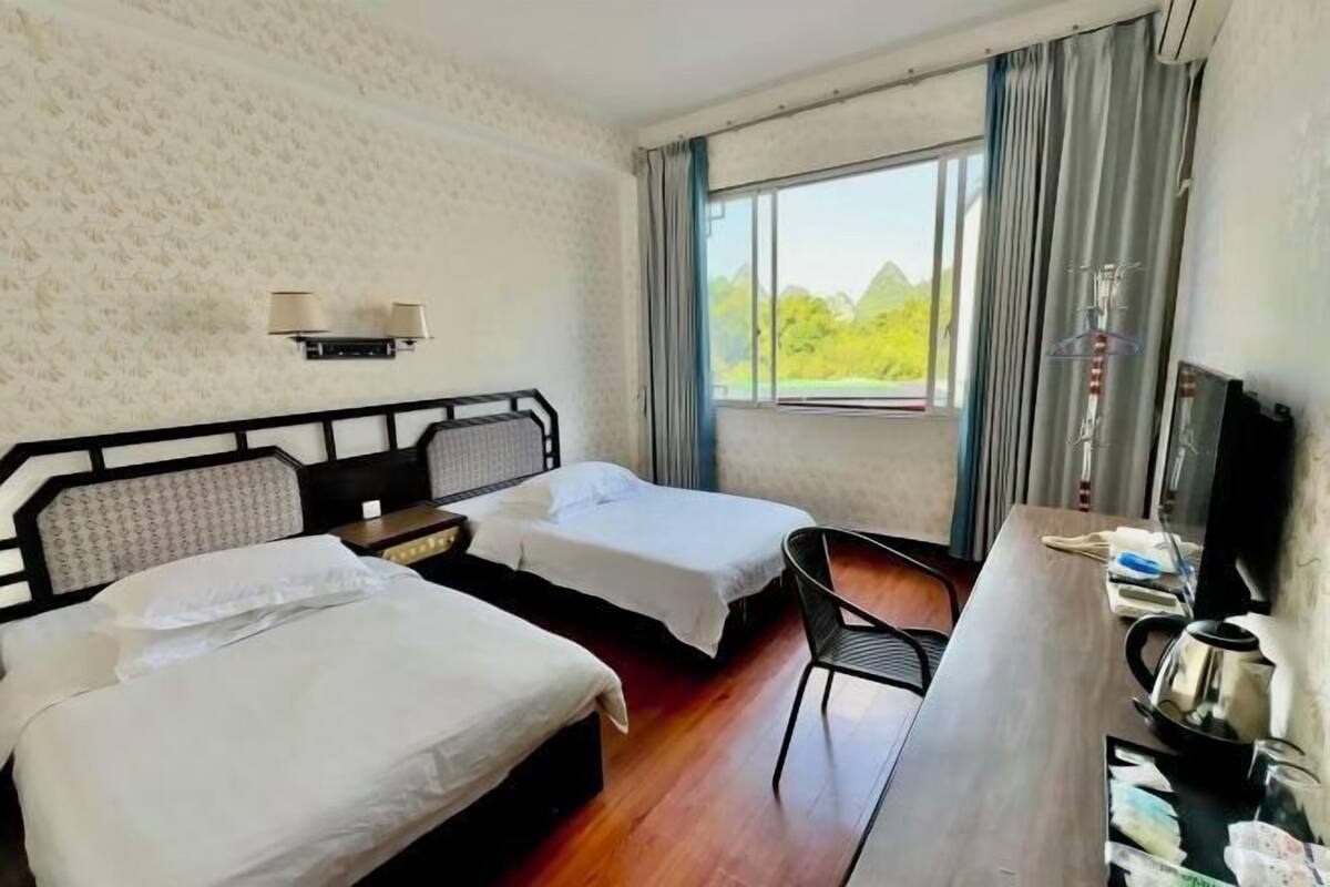 A hotel room with two beds, a desk, a chair, a television, and a window with a view of greenery.