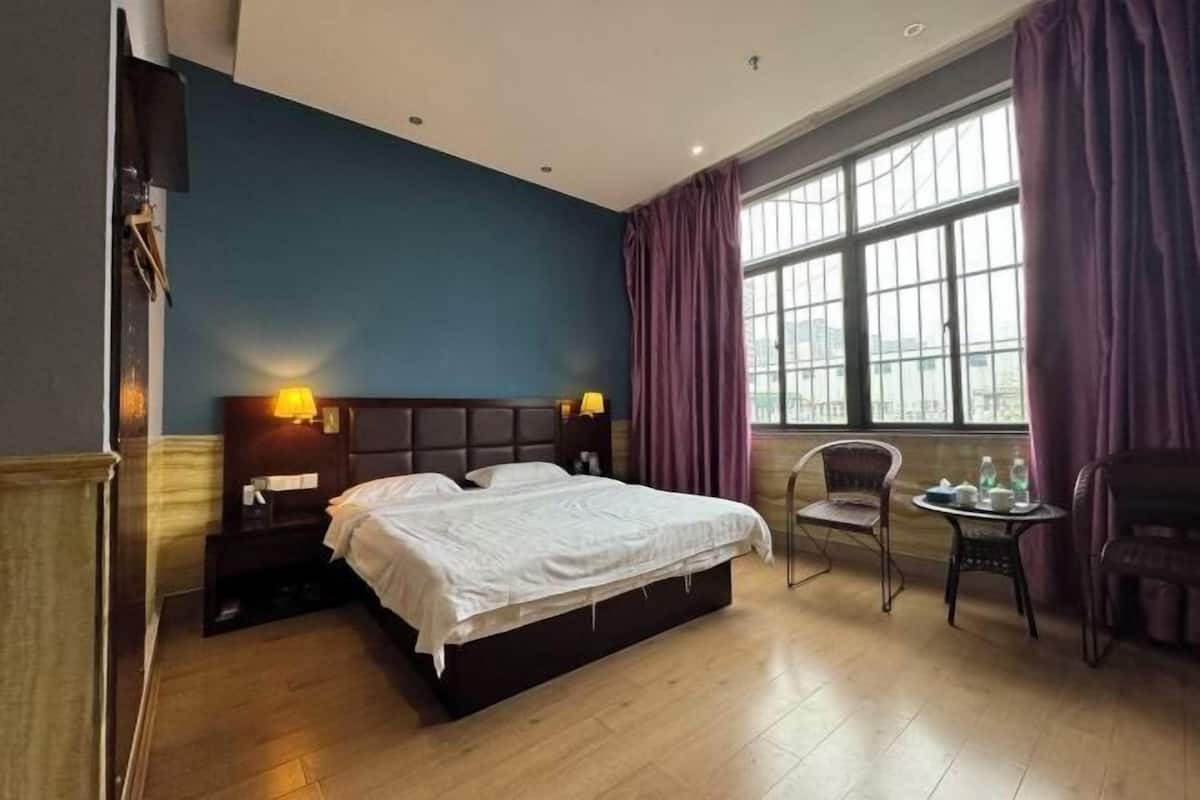 Quarto de hotel com uma cama grande, duas luminárias de cabeceira, uma pequena mesa com uma cadeira e uma janela com cortinas.