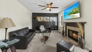 Ein Wohnzimmer mit Kamin, Deckenventilator, einem an der Wand montierten Fernseher und einem großen Strandbild.