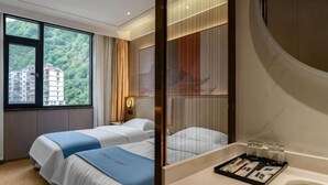 A hotel room with two beds, a large window with a view of buildings, a bathroom with a mirror, and a tray with toiletries. - New Beacon Shennongjia Villa (Shennongjia)