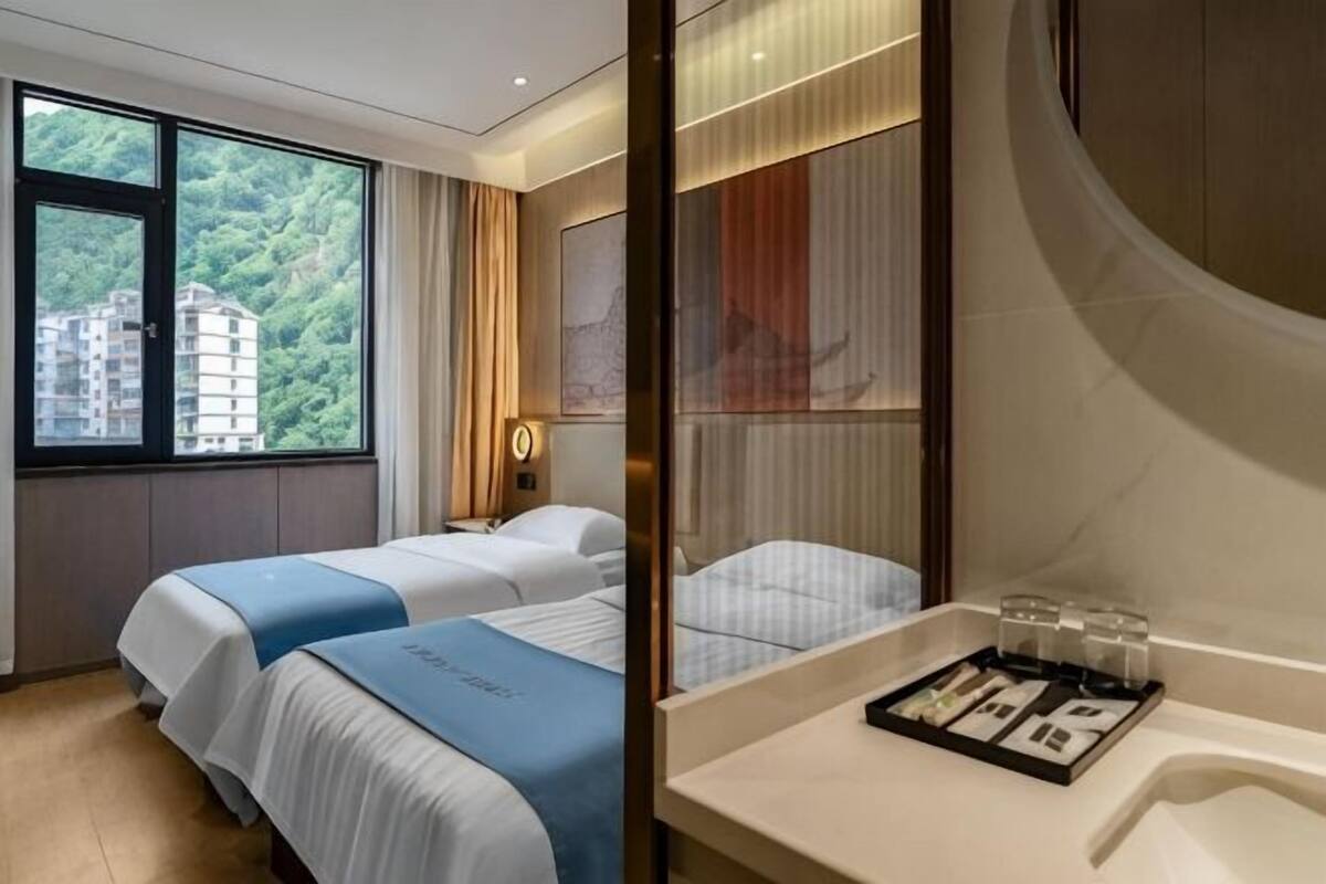 A hotel room with two beds, a large window with a view of buildings, a bathroom with a mirror, and a tray with toiletries.