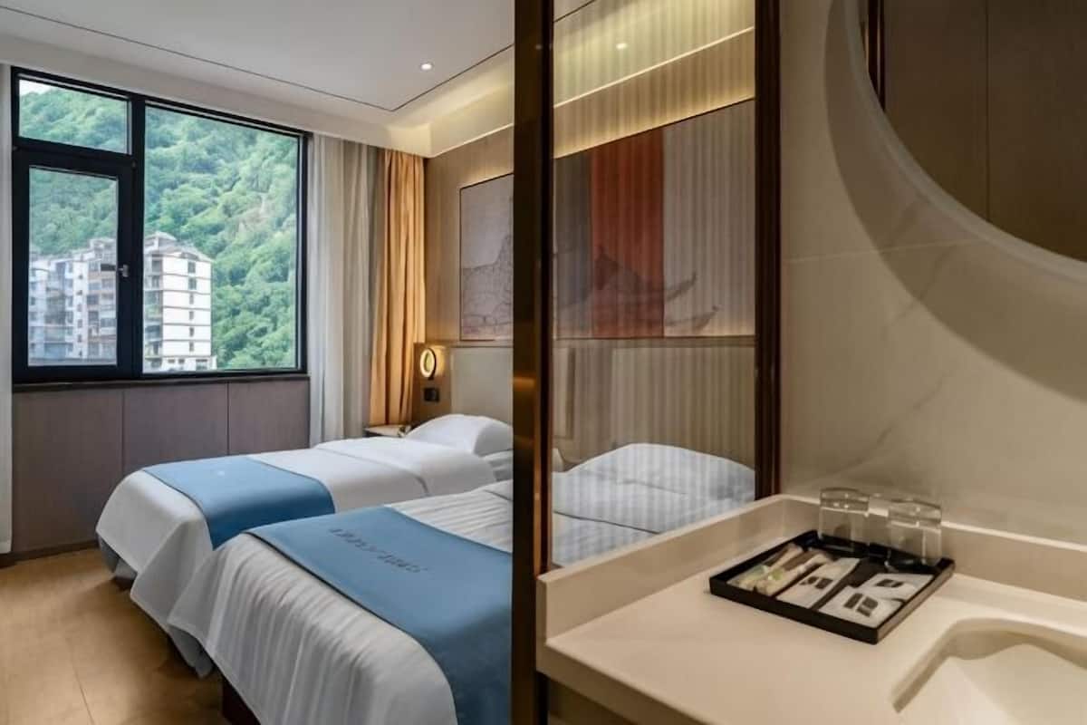 A hotel room with two beds, a large window with a view of buildings, a bathroom with a mirror, and a tray with toiletries.