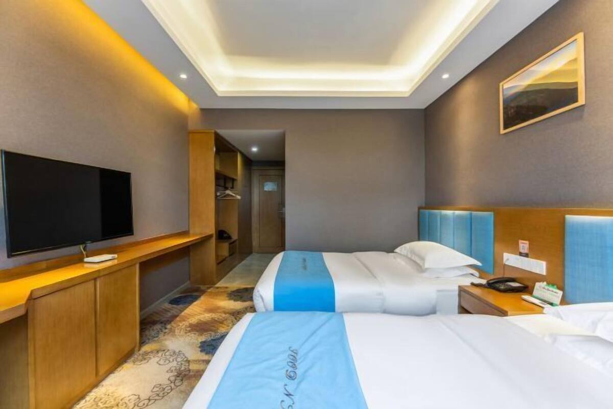 A hotel room with a flat-screen TV, a bed with a blue and white bedspread, a wooden headboard, and a small bedside table with a phone and a notepad.