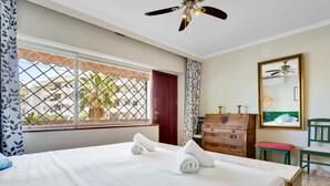 A hotel room with a bed, a dresser, a chair, a mirror, and a window with a view of buildings and palm trees.