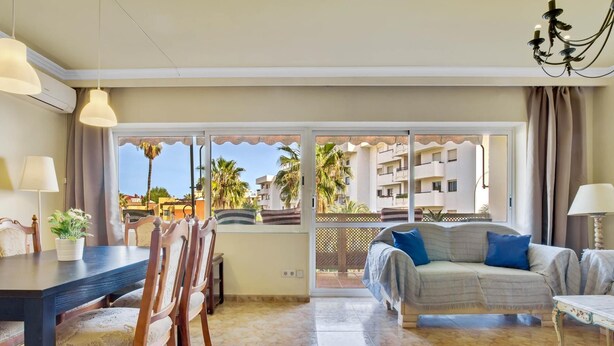 A spacious living area with a dining table, chairs, a sofa, and a balcony with a view of palm trees and buildings.
