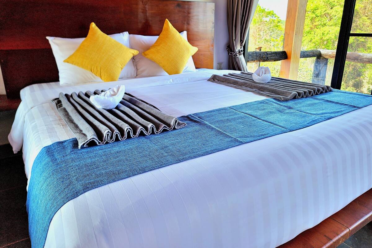 Cama bem arrumada com lençóis brancos, duas almofadas amarelas, um cobertor listrado em azul e branco e uma toalha listrada em castanho e branco.