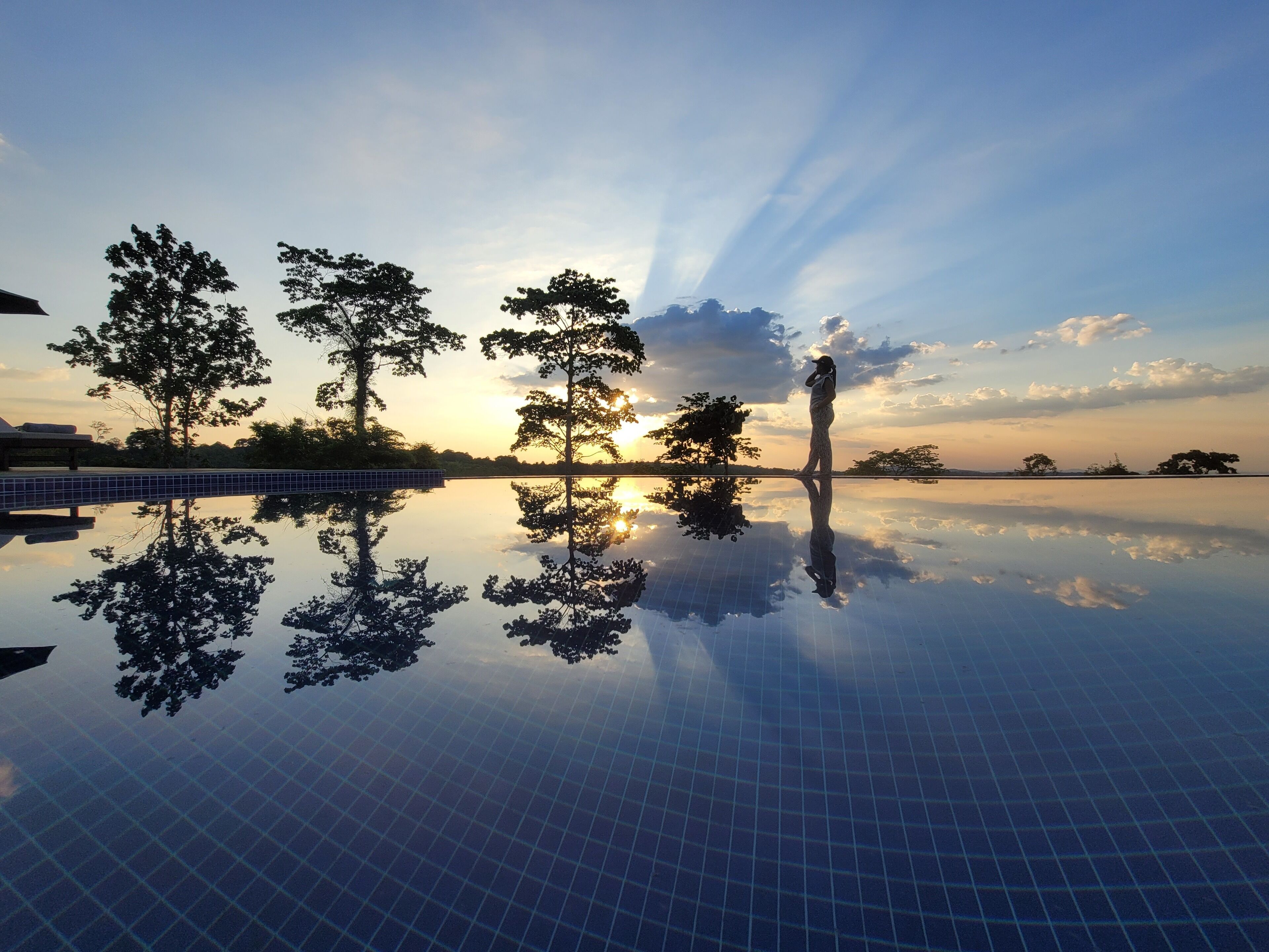 A serene outdoor pool area with a clear reflection of trees and the sky, surrounded by a tiled deck.