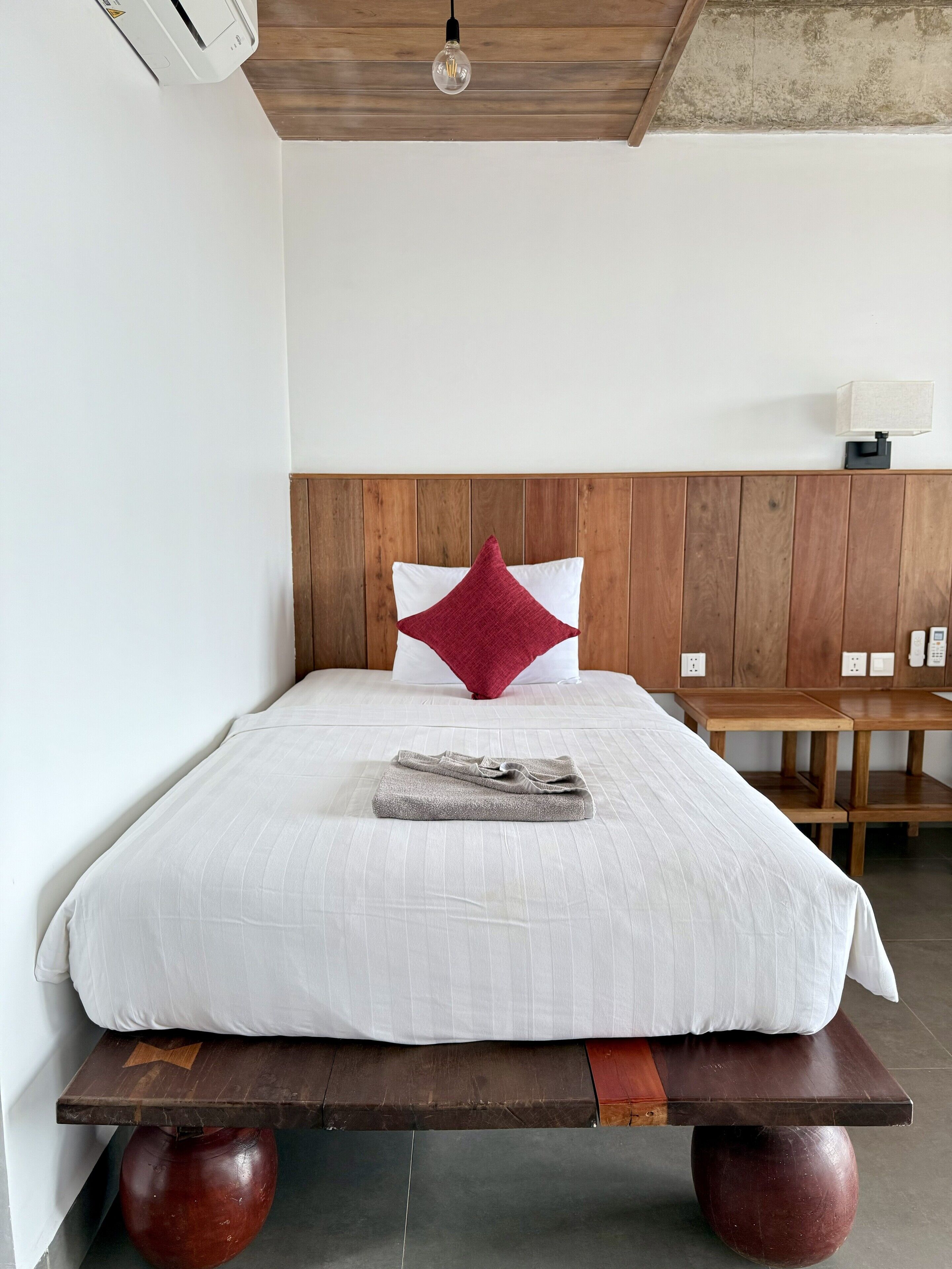 A neatly made bed with a white comforter and a red accent pillow, a wooden headboard, a bedside table, and a wall-mounted lamp.